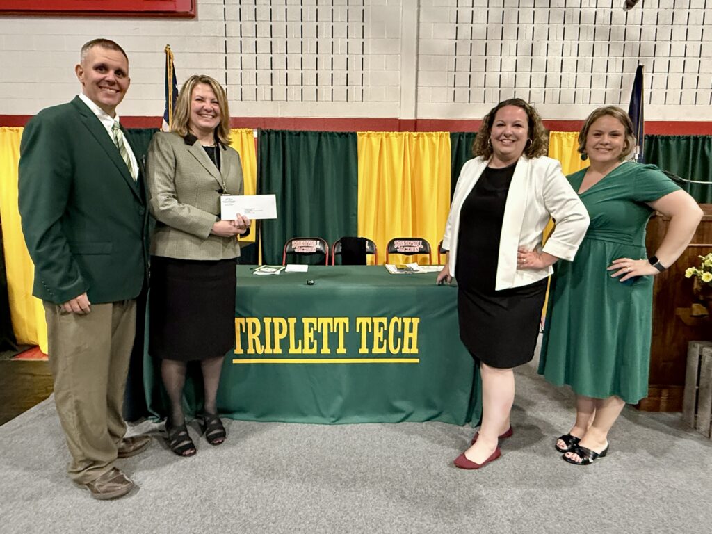 Four people stand together at a "Triplett Tech" event, indoors with colorful curtains and a table with a green tablecloth.